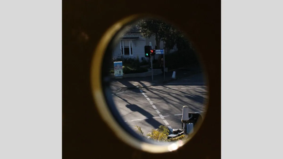 An urban street scene viewed through a small circular opening. The image frames a green traffic light, a pedestrian, and the corner of a house. Shadows fall across the road.