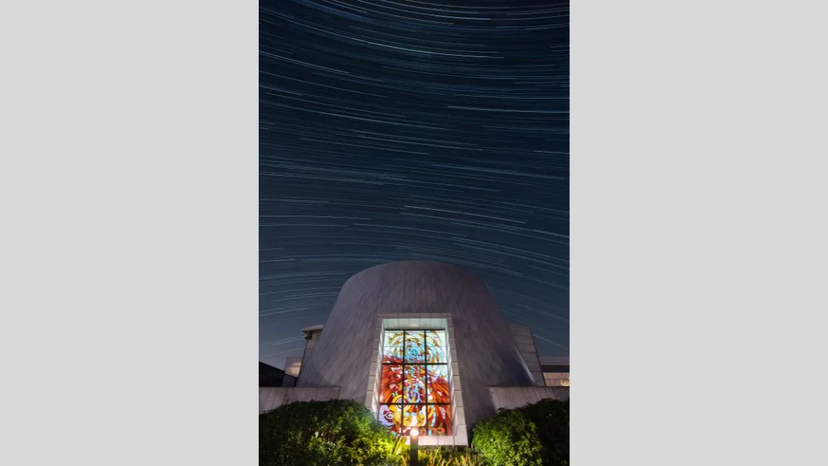 A modern white building with a large stained-glass window centered in the frame. The night sky above is captured in a long exposure, showing streaked star trails curving across the image.