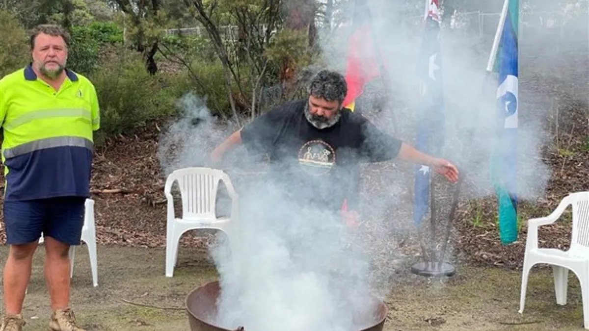 Smoking billowing out of a fire pit as an Aboriginal man conducts a smoking ceremony.