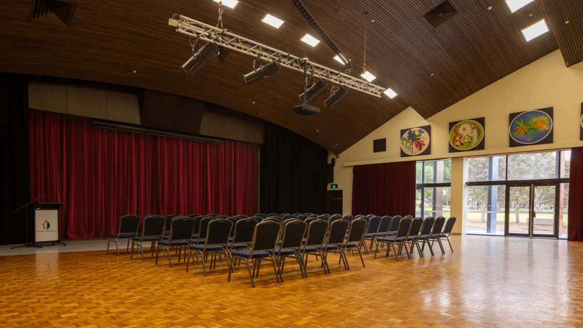 A room with rows of chairs facing a stage area with red curtains. Glass doors look out into a green area. The room has a wooden floor