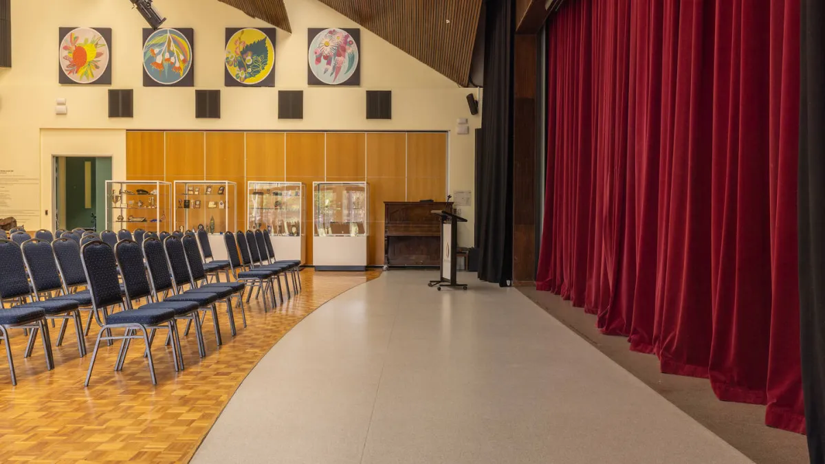 A room with rows of seats facing towards a staged area with podium and large red curtains. 