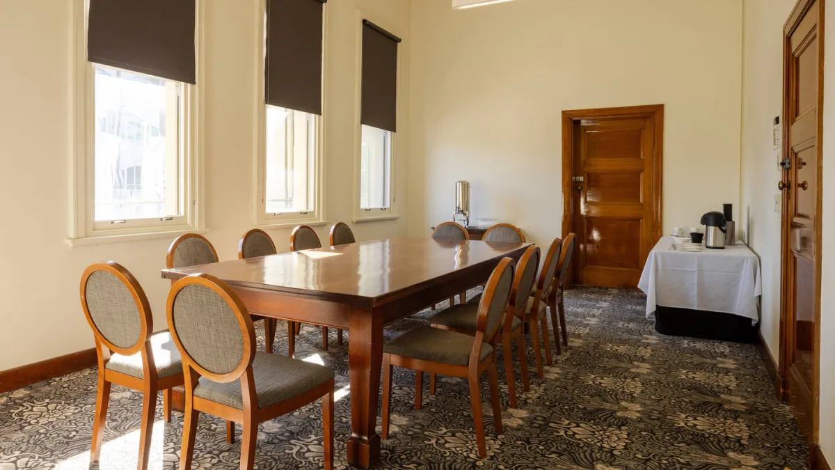 A meeting room with a large table with 12 chairs. A table with a kettle and cups is in the back corner. 