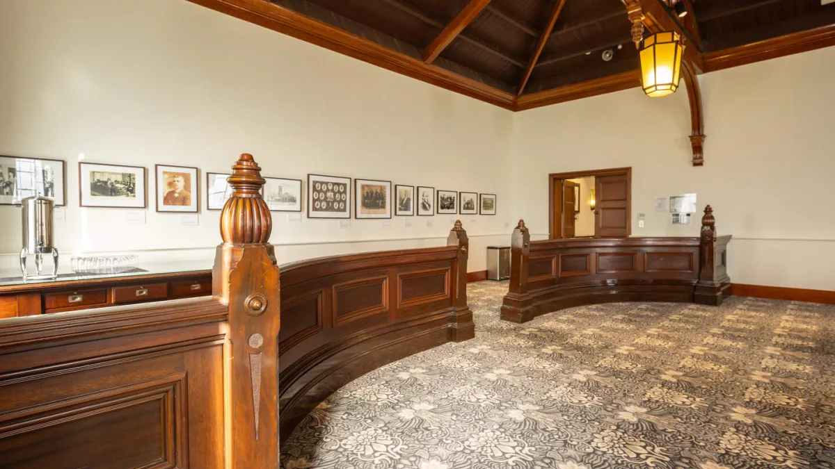 Interior of a historic building featuring a high, dark-wood ceiling, off-white walls lined with framed photographs, curved wood railings, and a patterned carpet.