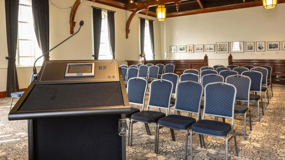 Interior of an elegant meeting room featuring rows of seating facing a podium, a decorative floor, and high ceilings with wooden beams.