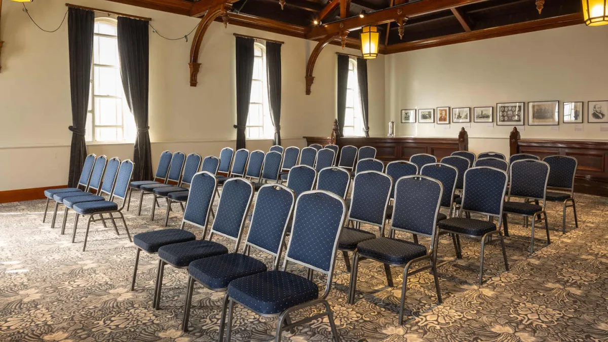 Interior of a room with rows of blue chairs facing forward. The room features patterned carpet and decorative ceiling.