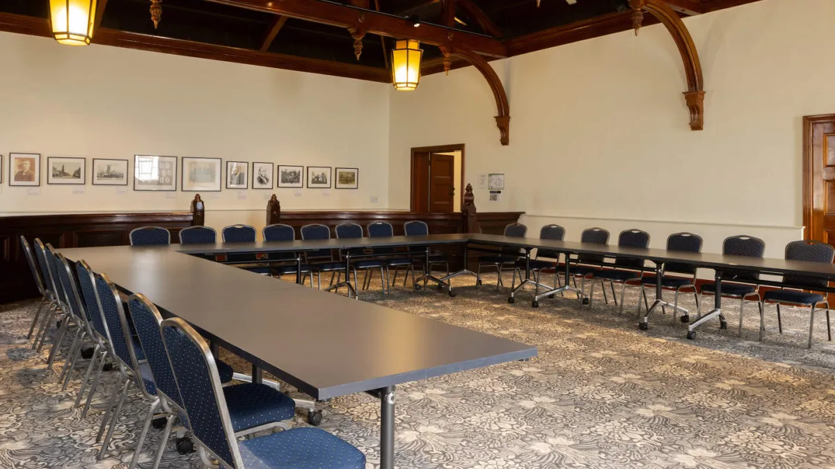 A large room with with long tables arranged in a U-shape. The room features dark wood ceiling, hanging lantern-style lights, and patterned carpet. 