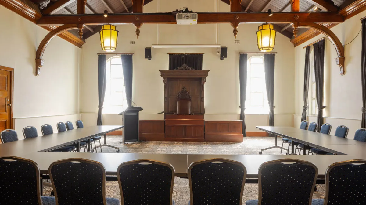 A conference room with long tables arranged in a U-shape, facing a raised platform with a wooden podium and antique-style furniture. The room has high, wood-beamed ceilings and arched details.