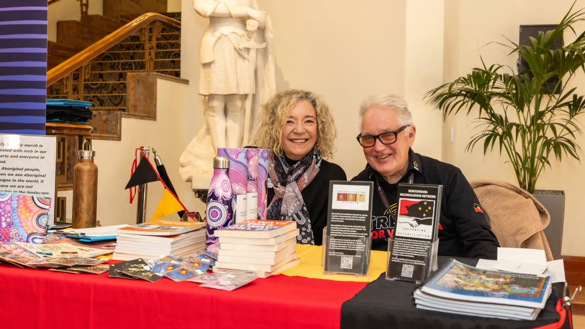 Two people smiling while seated behind a table covered with leaflets and brochures.
