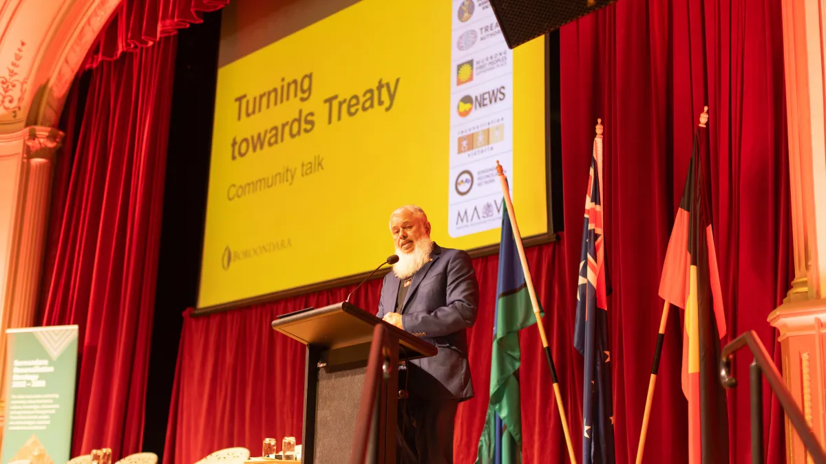 A man stands on stage behind a lectern, with a PowerPoint presentation displayed in the background.