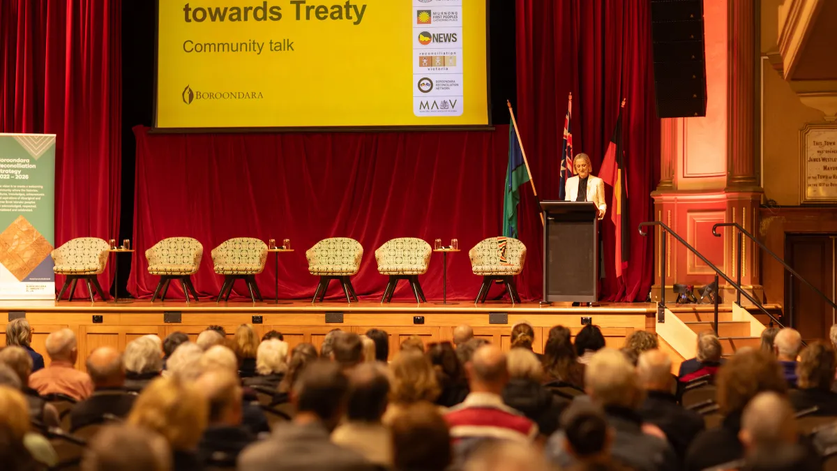 Audience members seated in a hall watch a person speaking on stage; several empty chairs on stage await a panel discussion.