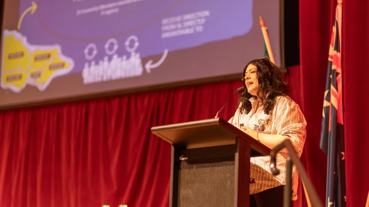 A woman stands behind a lectern on stage, presenting with a PowerPoint slide displayed in the background.