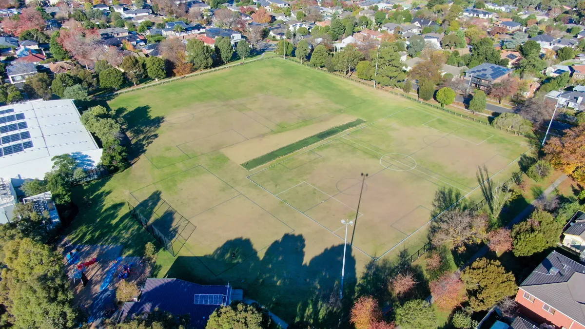 Aerial view of a retangular sportsground including a cricket pitch. It is surrounded by trees.