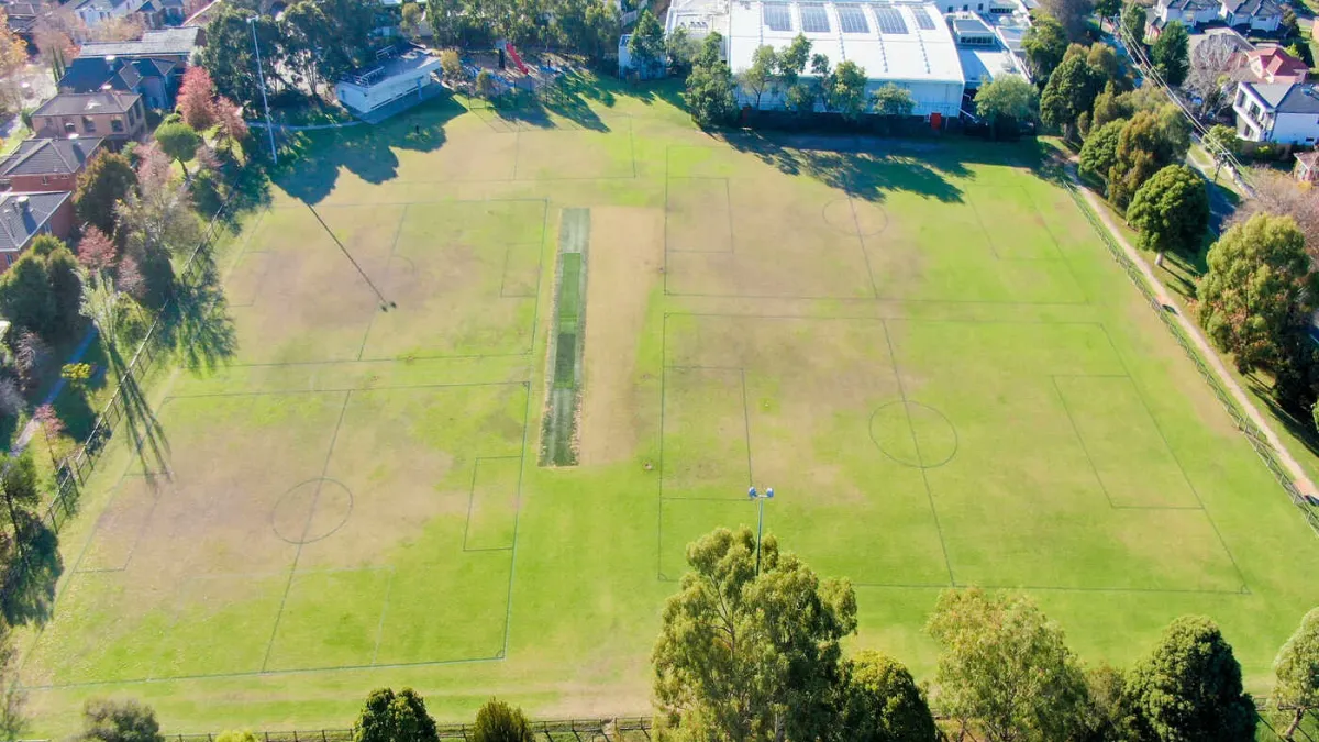 Aerial view of a retangular fenced sportsground including a cricket pitch. It is surrounded by trees, a pavilion and a playground