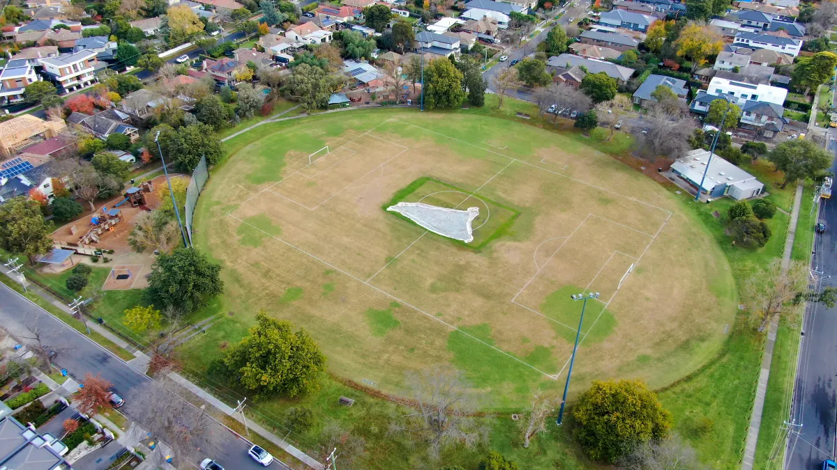 An aerial view of a oval sportsground. It is surrounded by a playground and houses.