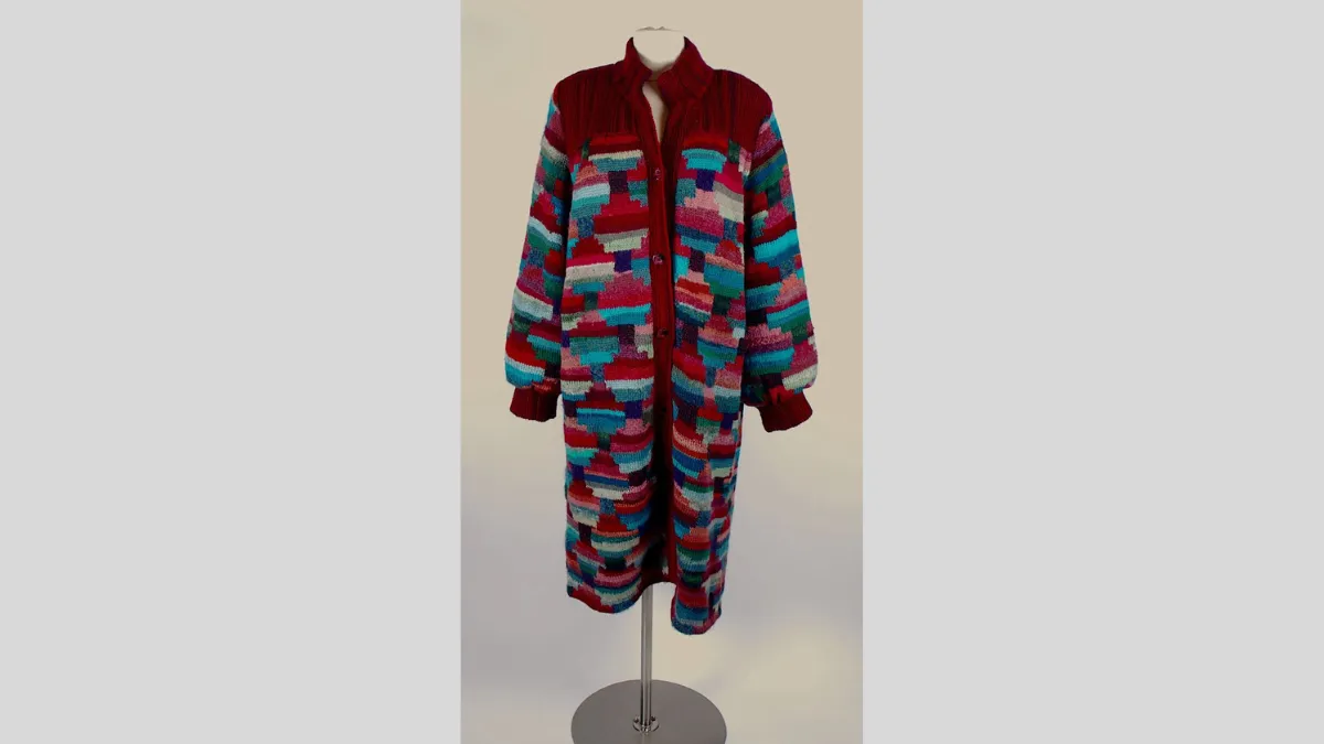 Photograph of a long-knitted coat dress displayed on a mannequin, featuring a button-down front and a high, ribbed collar. The garment has a red base with multicoloured block patterns in shades of teal, pink, purple, and grey. The cuffs are ribbed and gathered, and the coat has a loose, straight silhouette that extends to mid-calf length.