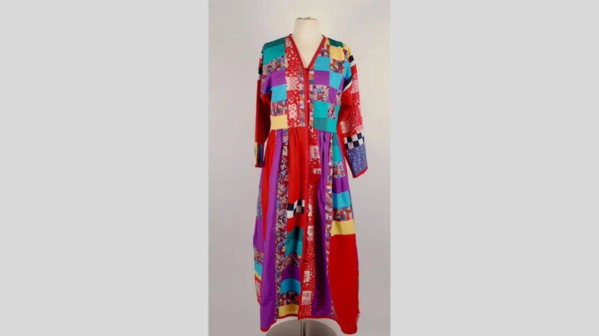 Photograph of a long-sleeved, ankle-length dressing gown displayed on a mannequin. The garment is constructed from a variety of colourful fabric patches, including florals, checks, polka dots, and solid colours like red, yellow, turquoise, purple, and black and white. The dress features a V-neckline, a buttoned bodice, and a gathered waist, creating a flared lower half.
