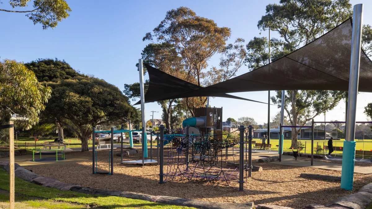 A small playground covered by a shade cloth with large trees behind it
