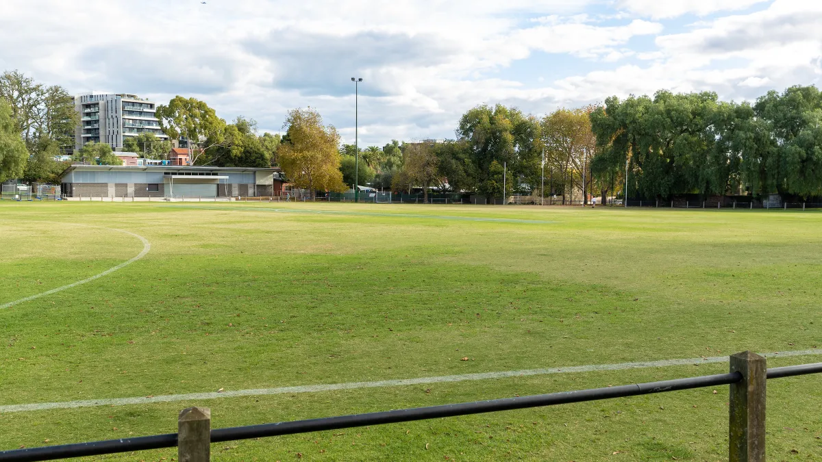 A playing field with a low fence. It is surrounded by trees. Footy goal posts and a small building are on the far side of the field