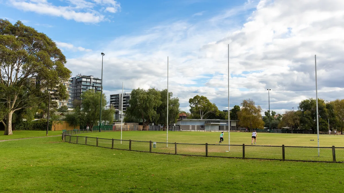 AFL oval with white goalposts, surrounded by a chain-link fence. Two people are running on the field. Trees line the edge, and apartment buildings are visible. 