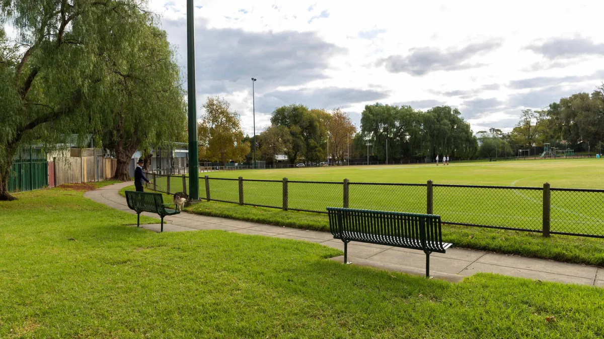 A park with benches and a grassy field. The park is fenced and it is surrounded by a walking path. 