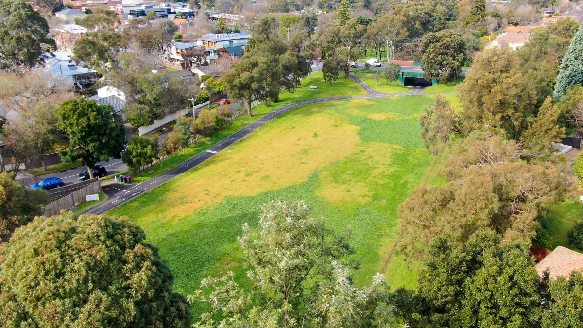 Aerial view of a grassy park area surrounded by trees. A path runs through the park leading to a tunnel under the road