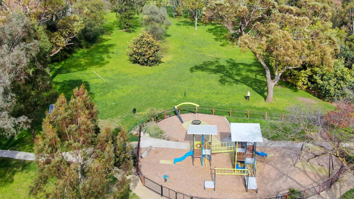 Aerial view of a park with a playground. The playground has slides, climbing structures, and a fence around it. There's a large grassy area beyond the playground with trees around the edge. 