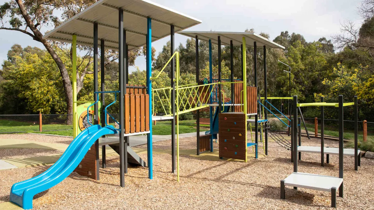 A playground with a mulch surface. It features 2 tower structures linked by a rope bridge, a slide, climbing wall and parallel bars. 