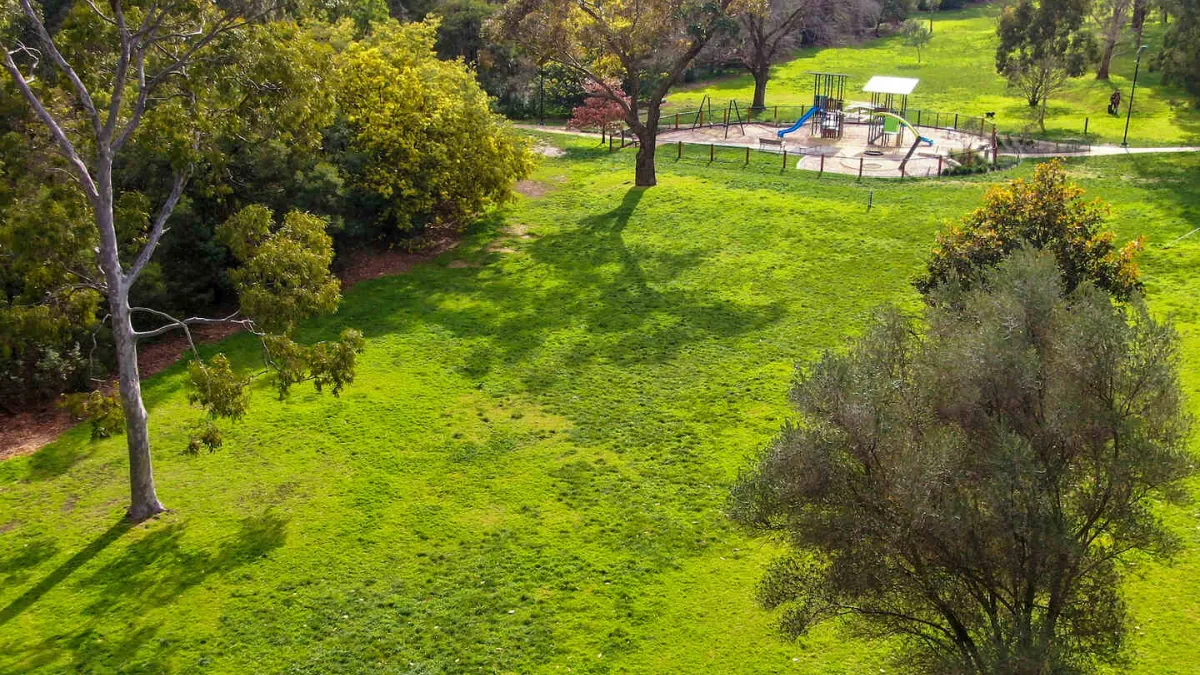 Aerial view of a large grassy area with trees. A fenced in playground is visible in the background. 