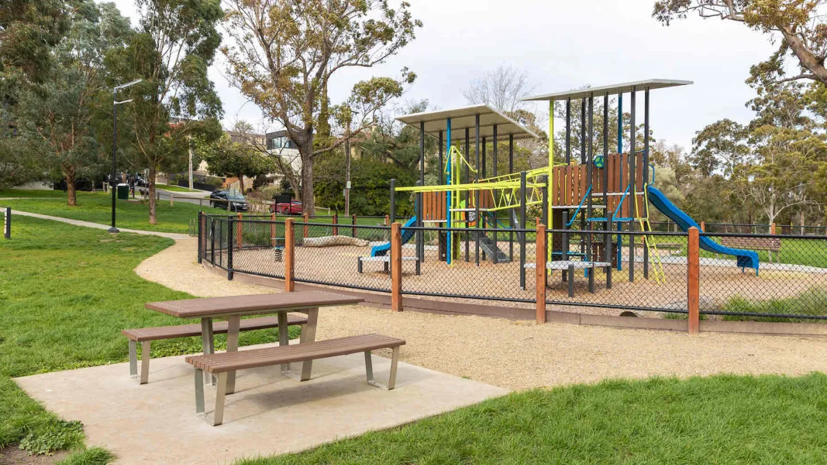 An outdoor playground with chain'link fencing featuring a climbing structurem and a slide. Outside the playground is a good picnic table. 