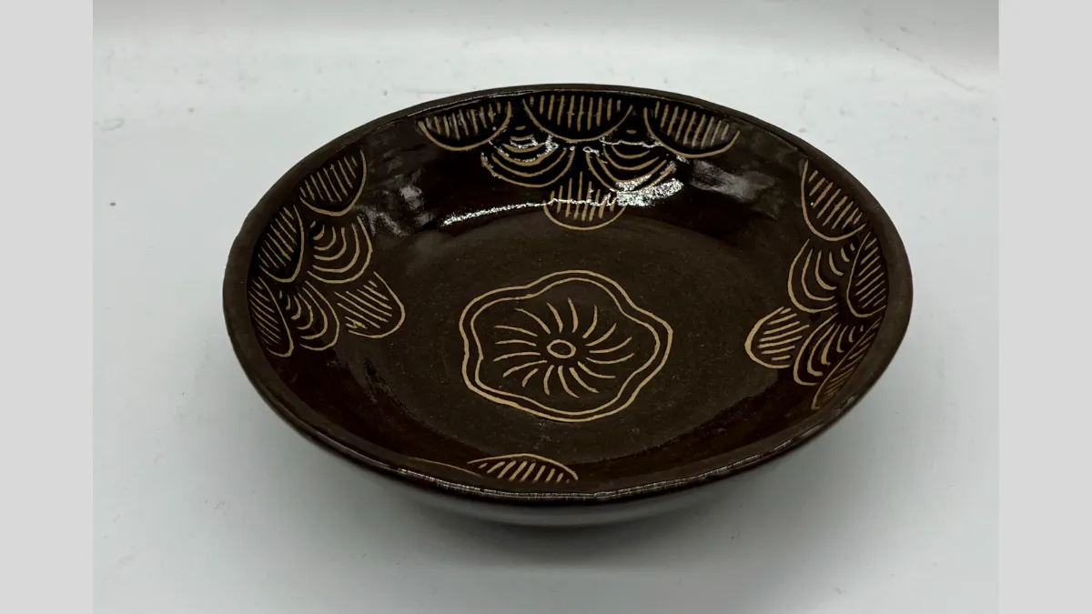 Photograph of a shallow, round ceramic dish with a dark brown glossy glaze. The interior surface is decorated with carved out floral and fan-like patterns in a lighter beige colour. 