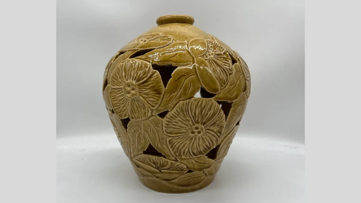 Photograph of a bulbous, light brown ceramic vase with a small opening at the top. The surface features carved floral and leaf motifs, with cut-out sections between them that create a lace-like design. The glaze is smooth and glossy.