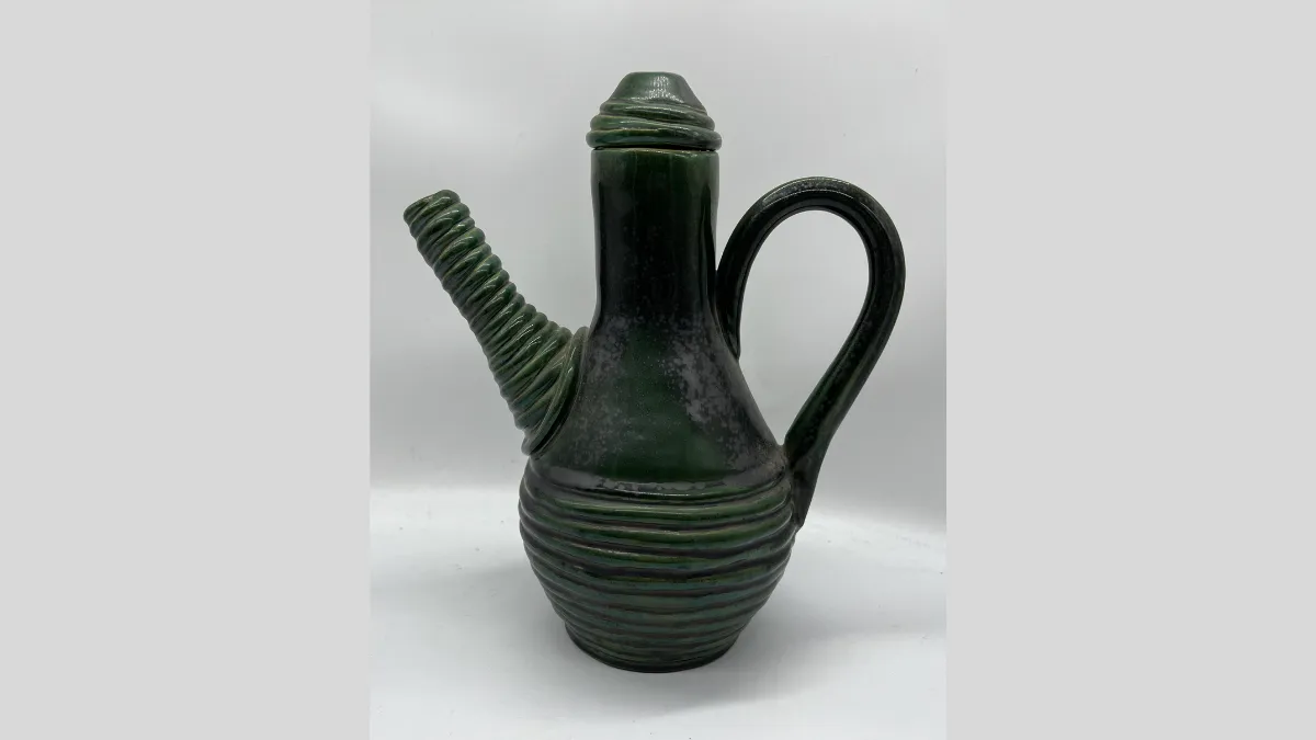 Photograph of a ceramic vessel with a deep green glaze, featuring a tall, narrow neck and a pointed lid. The spout is short and angled, with a ridged, spiral design. The body is rounded and decorated with evenly spaced horizontal ridges, and a single curved handle extends from the upper neck to the midsection.