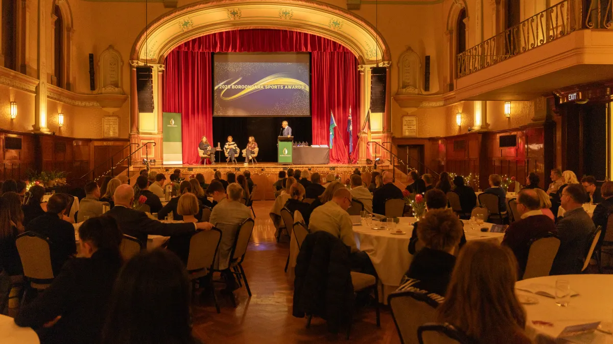 Banquet tables fill a fancy room, looking towards a stage where 3 people are seated and one is standing