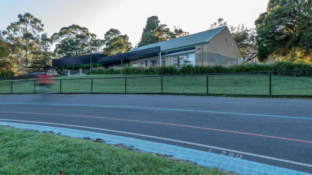 A velodrome track with blue and red line markets. In the background is short wire fence, and a building surrounded by trees.