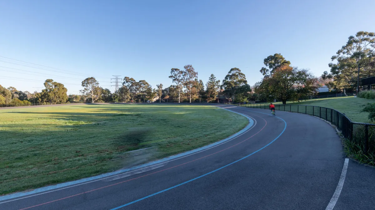 Outdoor scene featuring a cyclist on a curved running/cycling track. There is a green field in the centre , and trees in the background