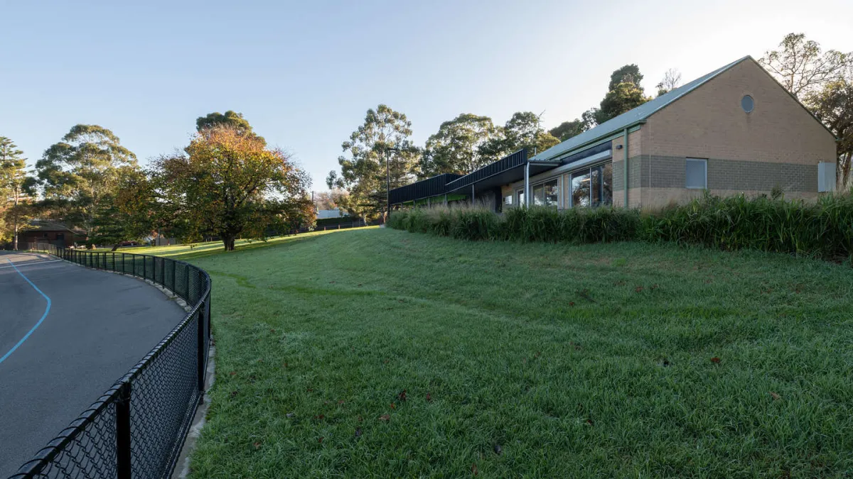 A grasssed area next to a fenced running/cycle track. There is a building in the background. 