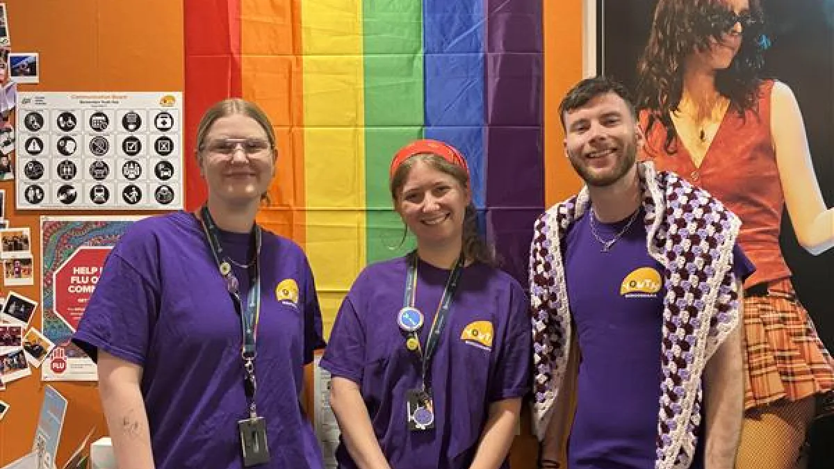 Three youn people in purple t-shirts standing in front of a pride flag hung on the wall behind them