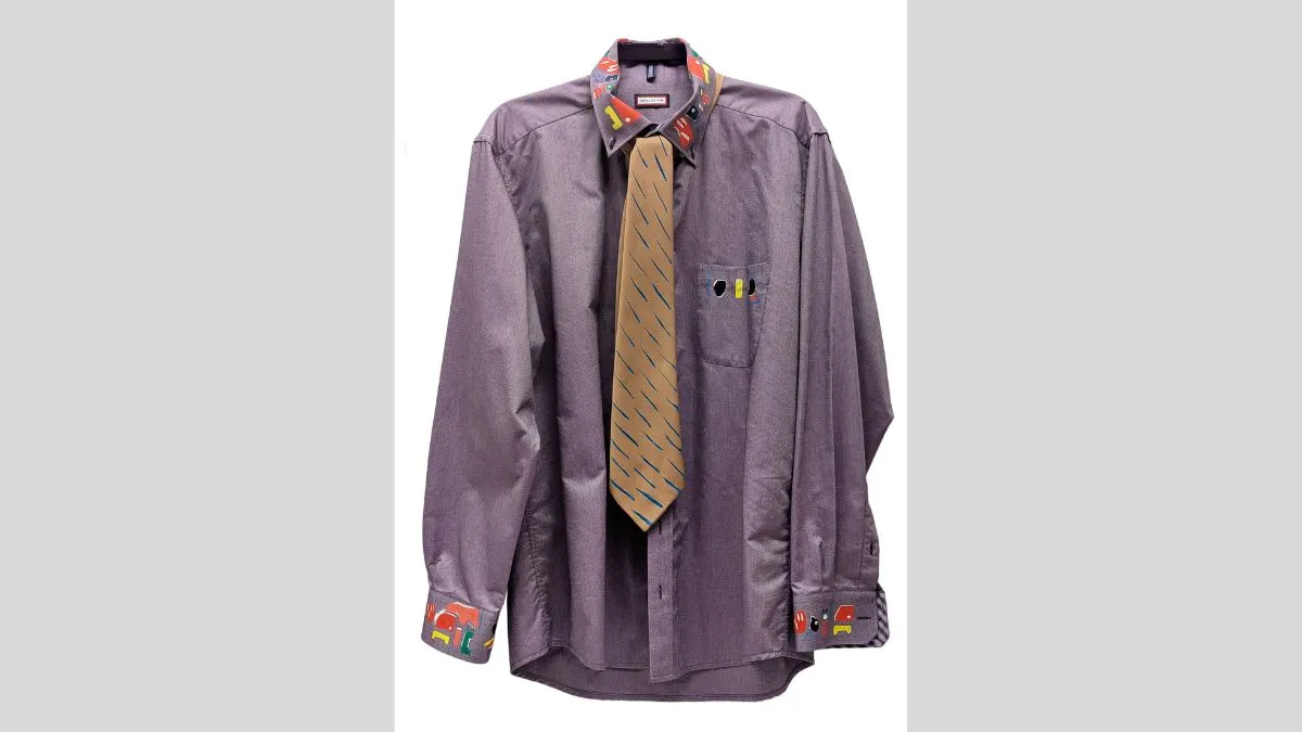 Photograph of a purple collared shirt. Collar, cuff of sleeves, and pocket are painted with colourful graphic shapes in red, yellow, blue, green and white. On top is a golden tie around the collar, painted with blue diagonal lines. 