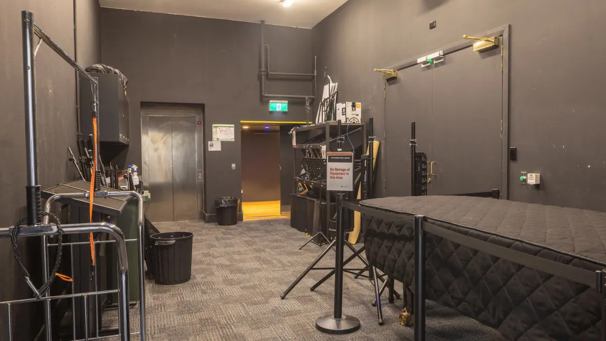 A storage room with dark gray walls and carpeted floor. An elevator is to the left. The room contains equipment such as a cart, lighting stands, and a covered table. A sign reads "No Storage of Equipment in this Area". A projector is mounted on the ceiling.