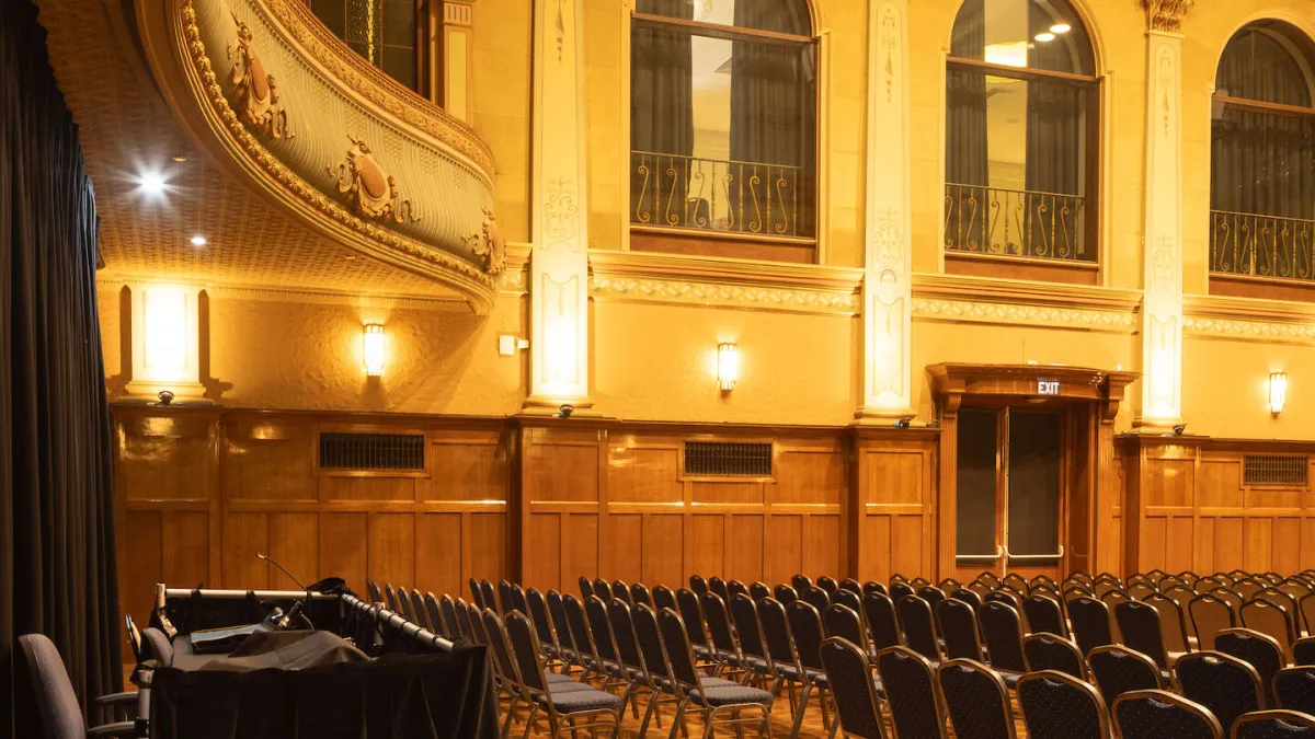 Auditorium interior with rows of chairs, stage, and decorative architectural details. A decorative balcony curves on the left side of the room.