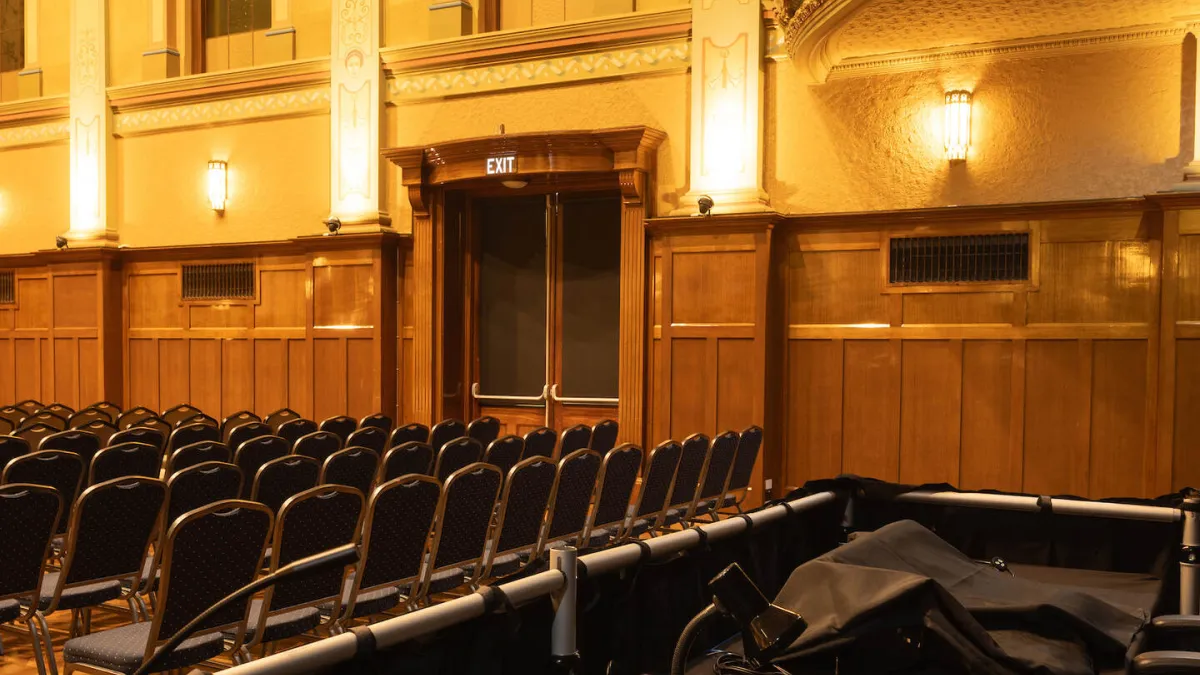 Stage and seating area inside a theater. A row of microphones sits on a stage platform. Numerous chairs with blue seats face the stage. The room has wood paneling and ornate architectural details, including an exit door with signage.