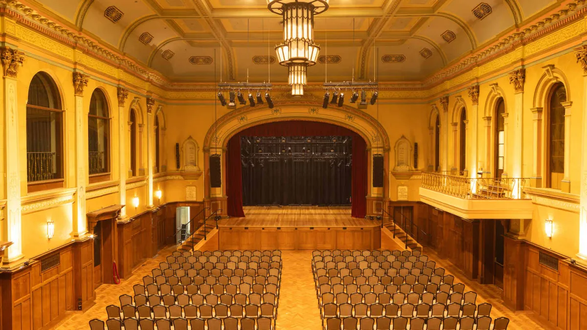 The spacious auditorium's interior includes a stage. Rows of chairs are set up in the main floor. There is a balcony on the right side and a large stage with red curtains at the far end. The room has ornate architectural details and is lit with chandeliers.