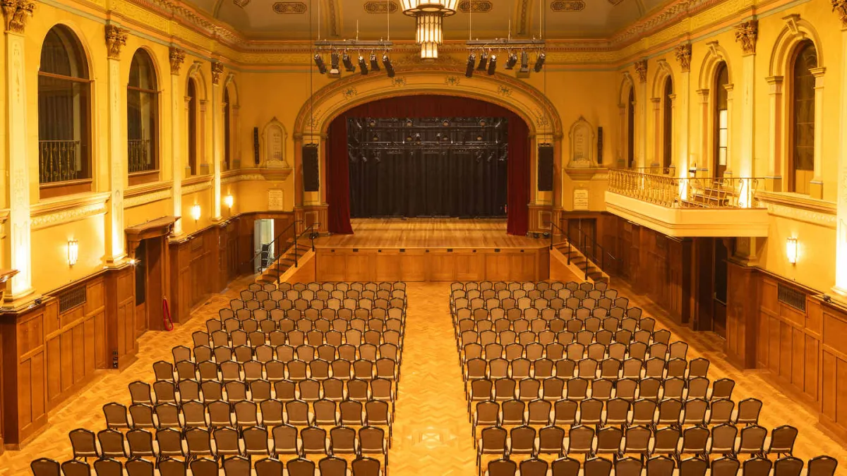 The spacious auditorium's interior includes a stage. Rows of chairs are set up in the main floor. There is a balcony on the right side and a large stage with red curtains at the far end. The room has ornate architectural details and is lit with chandeliers.