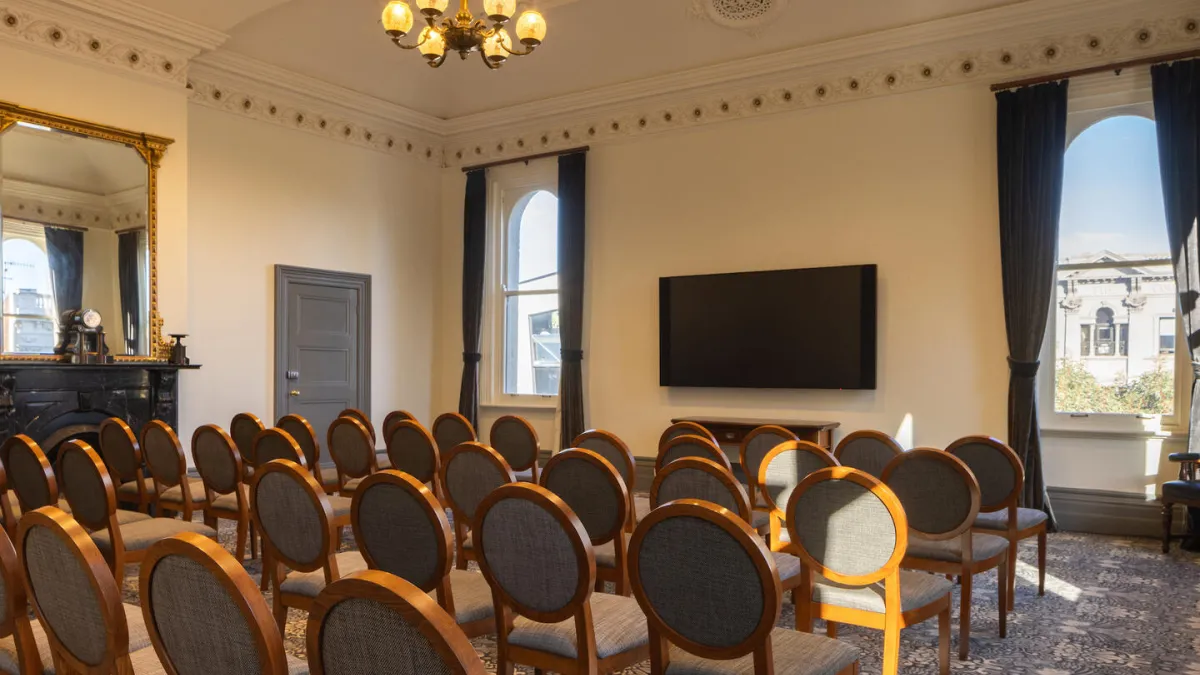 A meeting room featuring rows of wooden chairs with gray fabric seats, arranged in front of a large, wall-mounted television. Two windows flank the screen, draped with dark curtains. An ornate fireplace and mirror are visible to the left. The room has detailed ceiling molding and a chandelier.