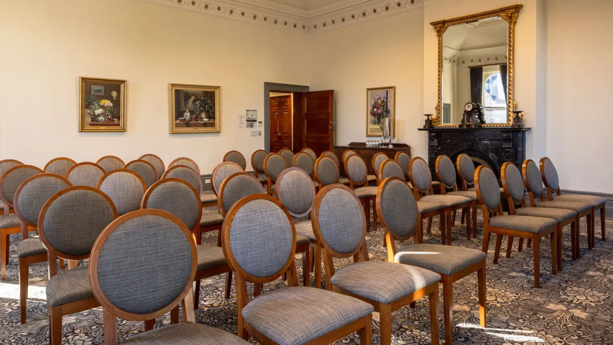 An room with rows of gray fabric chairs. A black fireplace topped with a large gold mirror is on the far wall. A chandelier hangs from the ceiling. The floor is covered in a patterned rug. A doorway is visible in the background.