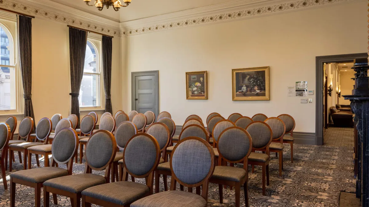 A room set up for an event, with rows of chairs facing forward. There are windows with drapes on the left, paintings on the wall, and a doorway on the right. The room has decorative molding and a patterned carpet.
