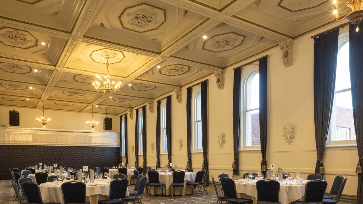 A banquet hall is arranged for an event. Round tables are set with white tablecloths, glasses, and silverware. Blue chairs encircle each table. The room boasts a high, ornate ceiling with chandeliers. Tall windows with dark curtains adorn the walls.