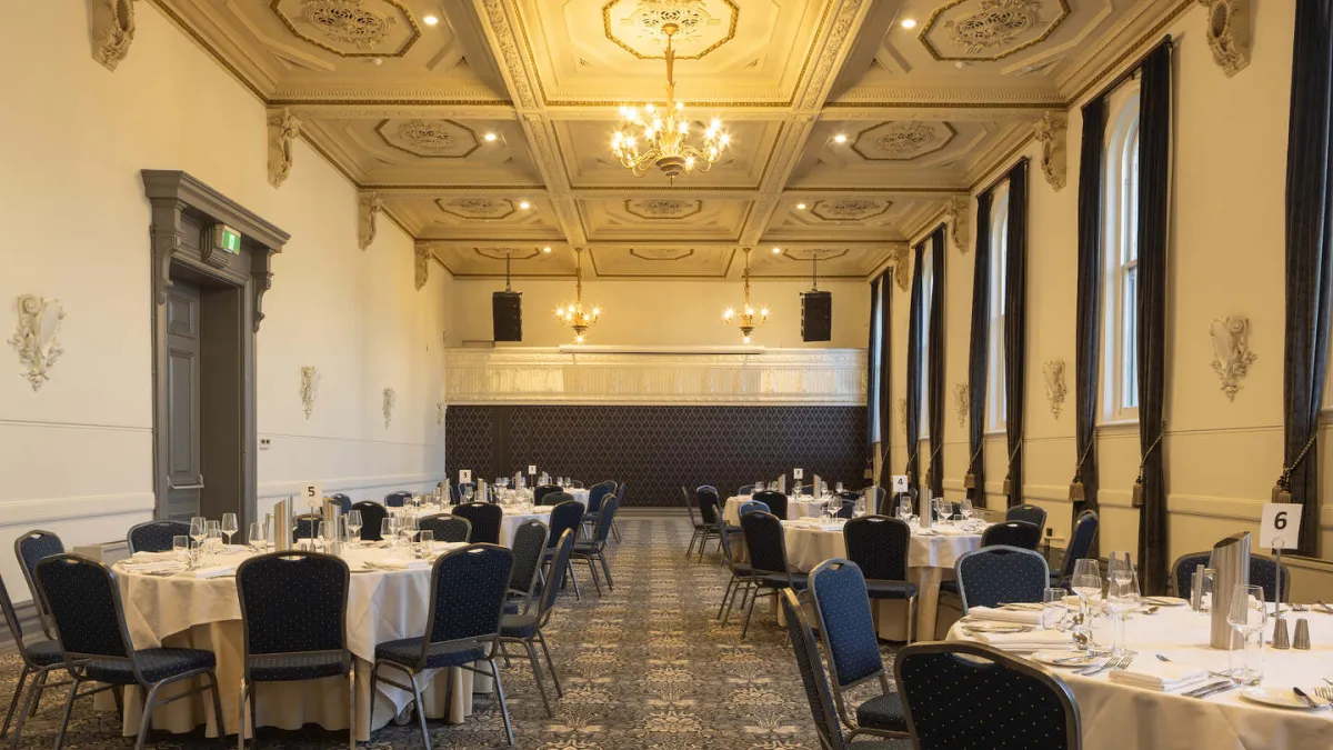 A ballroom with round tables arranged for a formal event. The room features ornate ceilings, large windows with dark curtains, and a floral patterned carpet. Each table is set with glasses, silverware, and a white tablecloth. Table numbers are displayed.