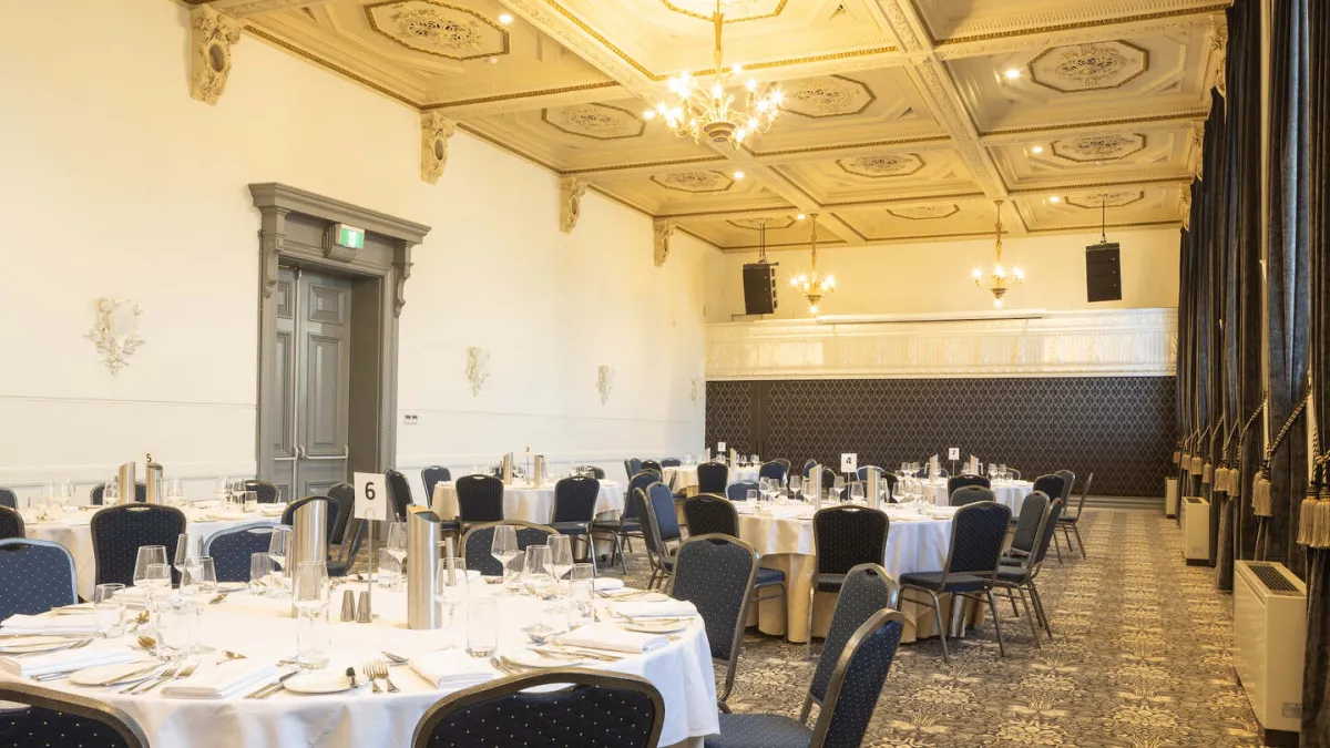A large room is set up for a formal dinner. Round tables with white tablecloths are arranged throughout the room, each set with silverware, plates, and glassware. Blue chairs with a gold polka dot pattern are around the tables. The room has high ceilings with decorative molding, ornate panels and several chandeliers. A raised stage area with speakers is in the background. Dark curtains are on the right. The floor has floral pattern carpet.