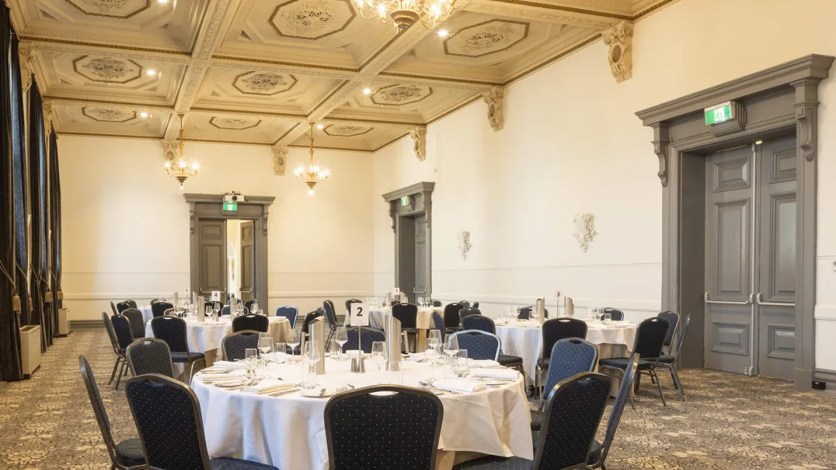A banquet hall is set up with round tables and chairs for an event. The ceiling is ornate with gold trim. Emergency exit doors are located around the edge of the room.